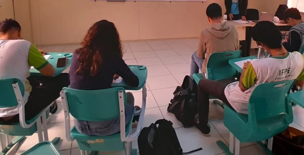Vista posterior de estudantes do IFPE Caruaru sentados em secretárias azul-turquesa numa sala de aula, realizando uma atividade de escrita individual